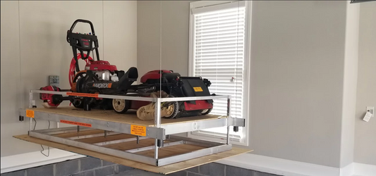 Aladdin storage lift raised near the garage ceiling holding a riding mower, pressure washer, and yard tools